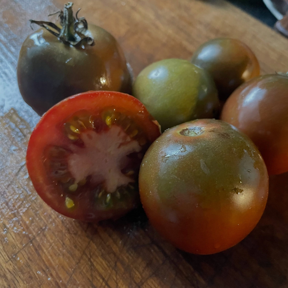 Tomates pequeños redondos con hombros verde oscuro y marrones sobre una tabla de madera, uno cortado por la mitad mostrando pulpa roja con gel verde alrededor de las semillas.