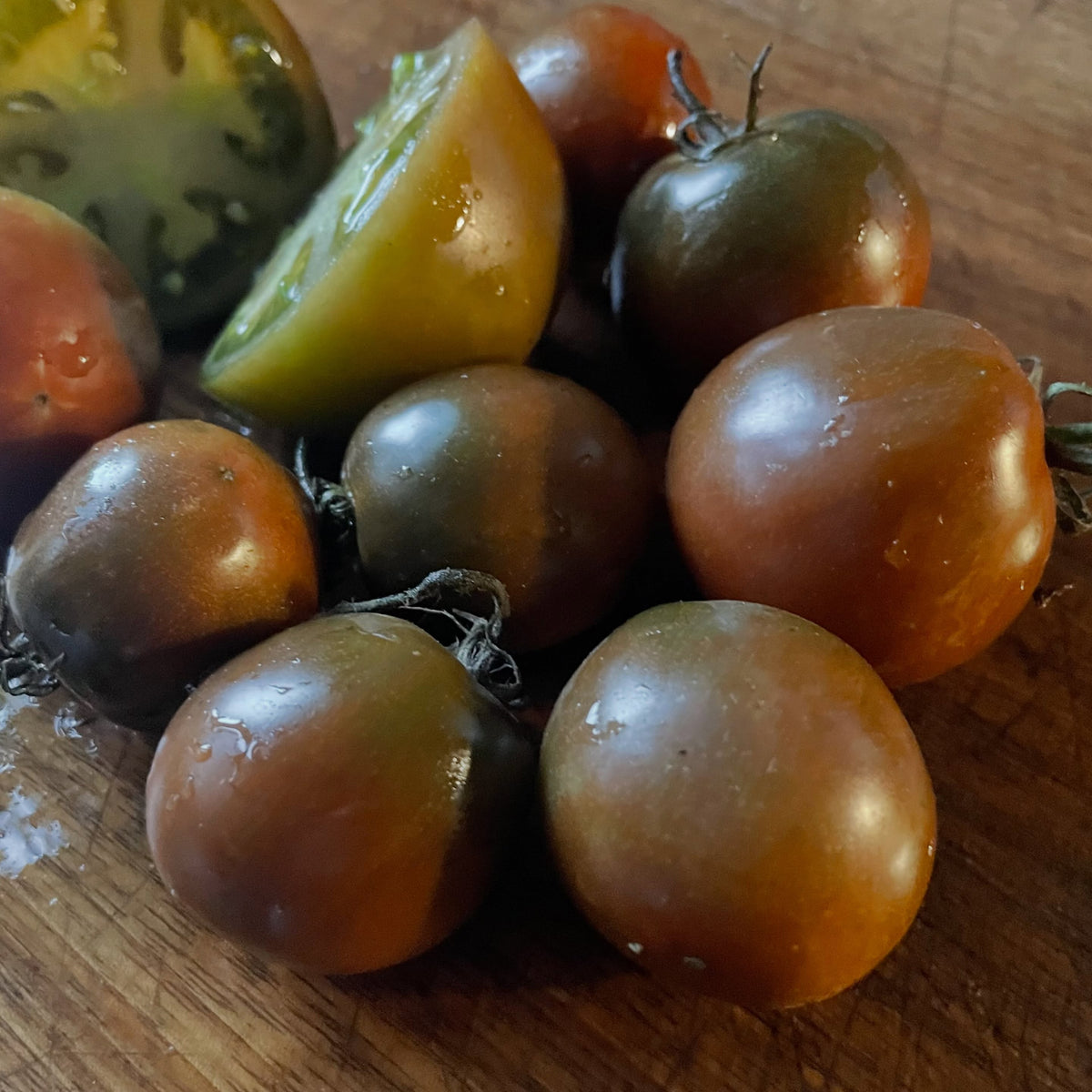 Un montón de tomates pequeños marrones y verdes sobre una tabla de madera, con dos trozos cortados que muestran pulpa verde y amarilla.